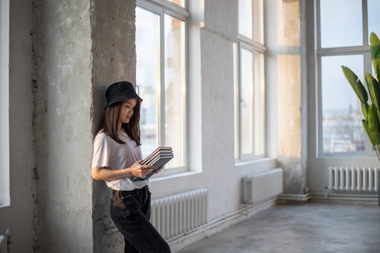 Young Woman In Hat Studying Books And Leaning On Wall