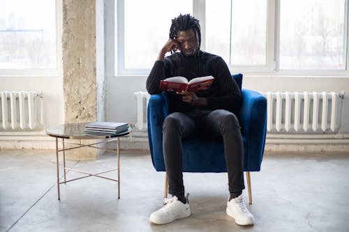 Free Full body of young African American male reading book in comfortable armchair near table with literature in hall in daytime Stock Photo