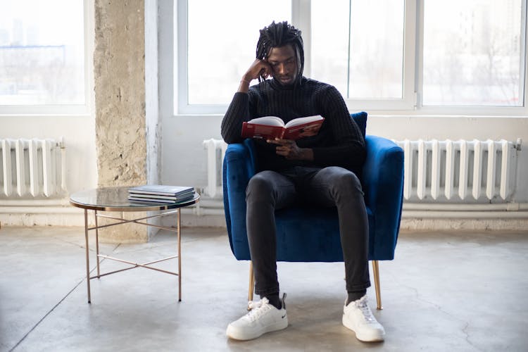 Focused Black Man Studying Book In Armchair Near Table