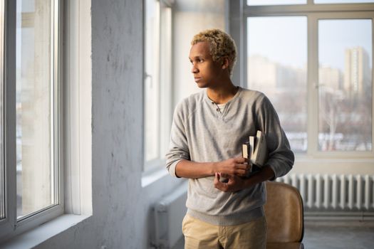 Thoughtful African American male in casual outfit with books looking away near windows in university