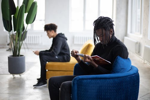 Thoughtful African American man with braids sitting on blue armchair and reading magazine against pensive black guy browsing smartphone sitting on couch in light spacious studio in daytime