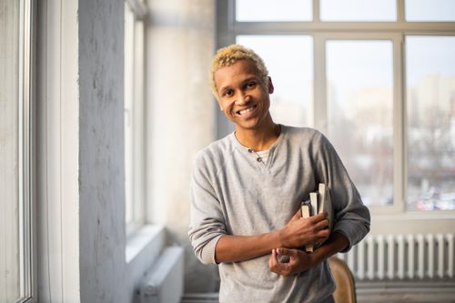 Free Cheerful African American male with light hair in casual clothes standing with books in hand in light spacious room and looking at camera Stock Photo