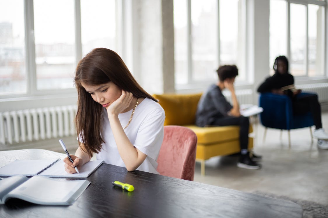 Free Concentrated young female student sitting at table with books and writing synopsis against blurred men communicating in light workspace in daytime Stock Photo