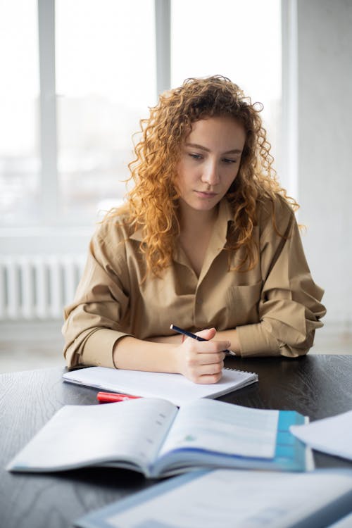 Concentrated young female with curly hair in casual shirt sitting at table with books and taking notes in notebook while studying in light workspace against window in daytime