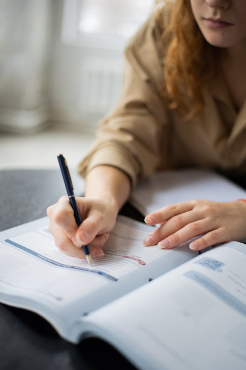 Free Unrecognizable female student with pen taking notes in book while sitting at table during studying in room on blurred background Stock Photo