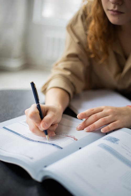 Unrecognizable female student with pen taking notes in book while sitting at table during studying in room on blurred background