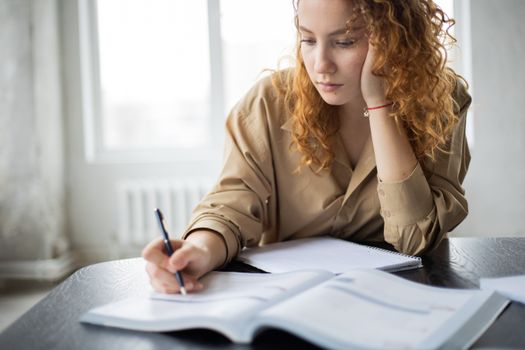 Diligent female student with copybook leaning on hand and taking notes in textbook at wooden table while studying in room