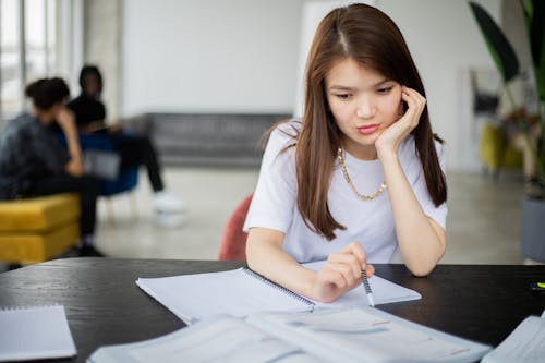 Wistful Asian female with pen leaning on hand and sitting at table with copybook during studies in room with classmates on blurred background