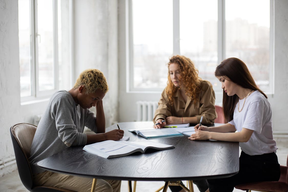 Free Group of focused multiracial classmates taking notes in notebooks while collaborating during preparation for lesson at wooden table with textbook Stock Photo