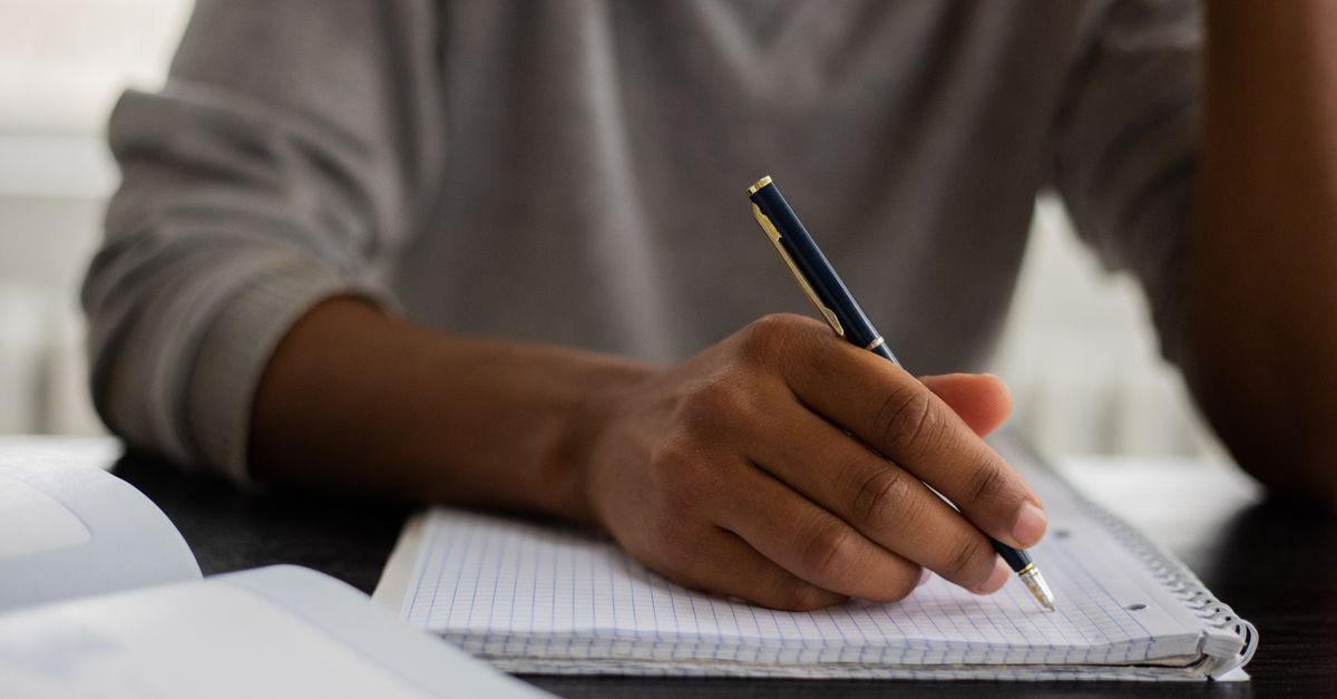 Unrecognizable African American male with pen taking notes in workbook while sitting at table with textbook on blurred background during studying