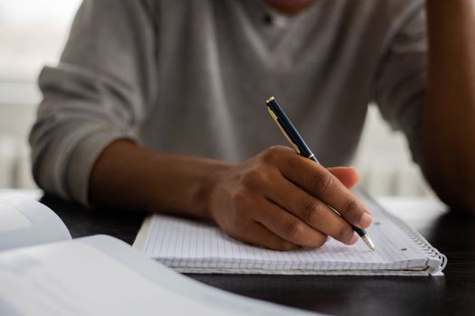 Unrecognizable African American male with pen taking notes in workbook while sitting at table with textbook on blurred background during studying