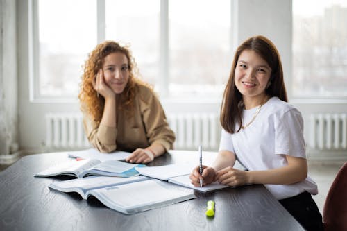 Smiling multiethnic female classmates looking at camera and taking notes in notebook while sitting at table with textbook and preparing for seminar