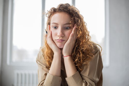 Free Pensive female student with hands on cheeks looking down while sitting in light room on blurred background while deciding task in frustration Stock Photo
