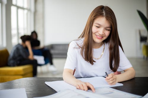 Smiling Asian female with pen and copybook reading textbook at table during lesson in room with classmates on blurred background