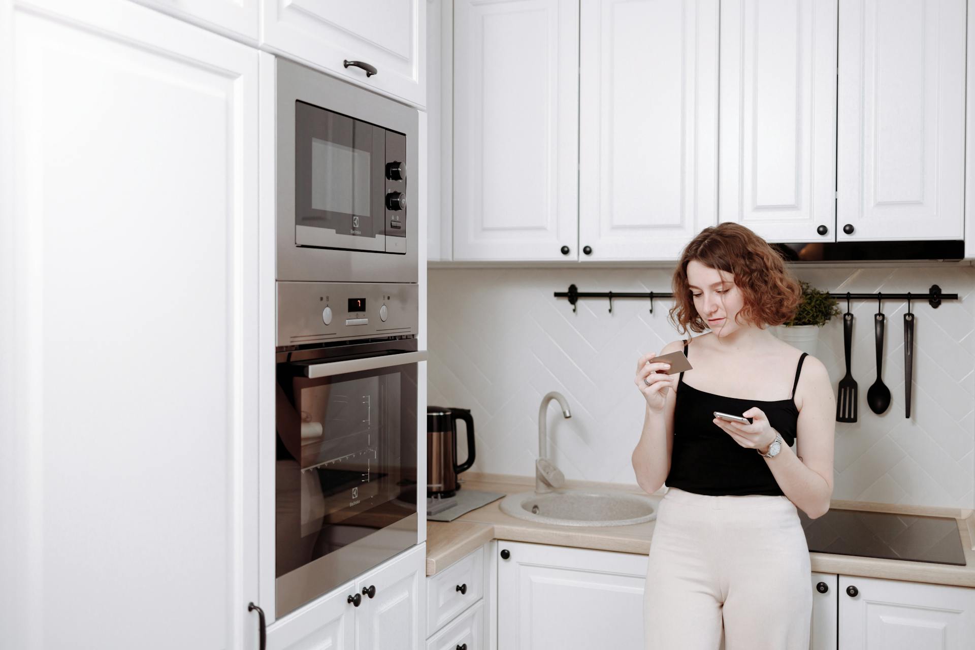 Young woman using phone and credit card for online shopping in stylish kitchen.