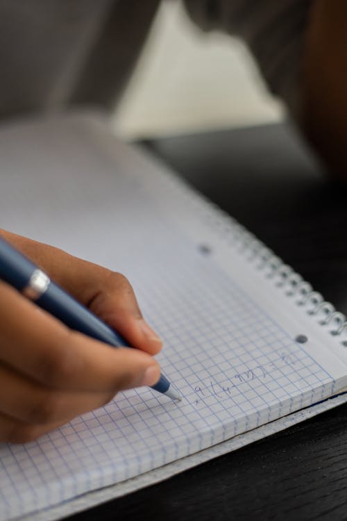 Unrecognizable African American person with pen taking notes in notepad while doing exercise in classroom during lesson on blurred background