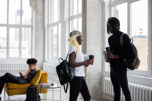 Free Side view of African American students with backpacks and takeaway hot drinks communicating and looking at each other while standing with copybooks in hands in classroom with classmate on couch Stock Photo