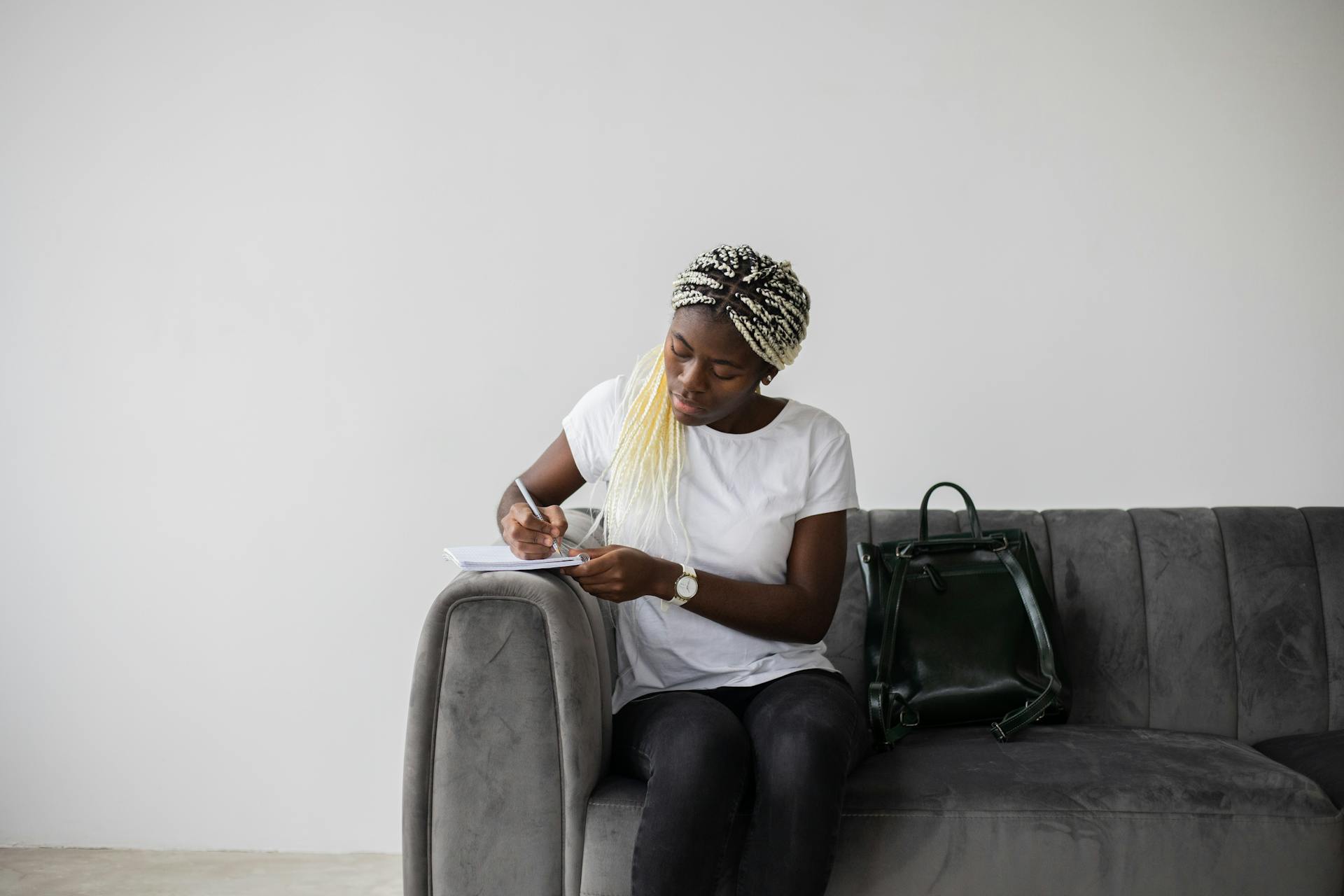 Focused African American female student with Afro braids taking notes in copybook while solving task on couch with backpack in room