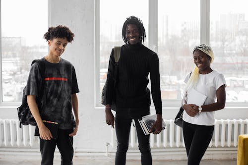 Group of smiling African American students with backpacks looking at camera while standing with copybooks in hands during lesson in classroom