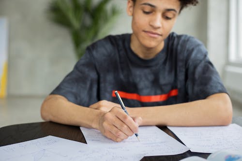 Crop African American male student taking notes on piece of paper at table while studying subject in classroom on blurred background