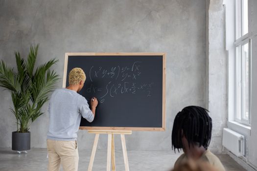 Unrecognizable African American male student writing mathematical formulas on chalkboard while studying in classroom with faceless black classmate during lesson