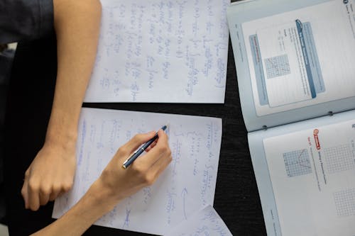 Top view of anonymous person with pen taking notes on piece of paper while solving equations in classroom during lesson