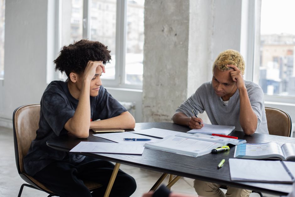 Focused African American male students with pens and copybooks sitting at table with textbooks while studying together in classroom during lesson Focused African American male students with pens and copybooks sitting at table with textbooks while studying together in classroom during lesson