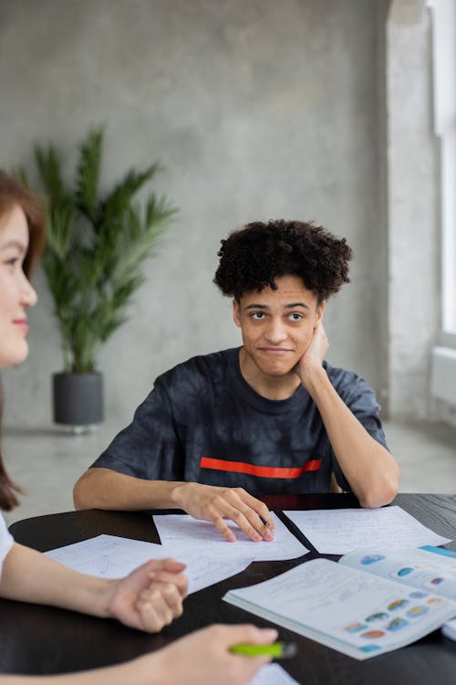Free African American male student sitting at table with books and papers and solving task during lesson in classroom with unrecognizable classmate Stock Photo