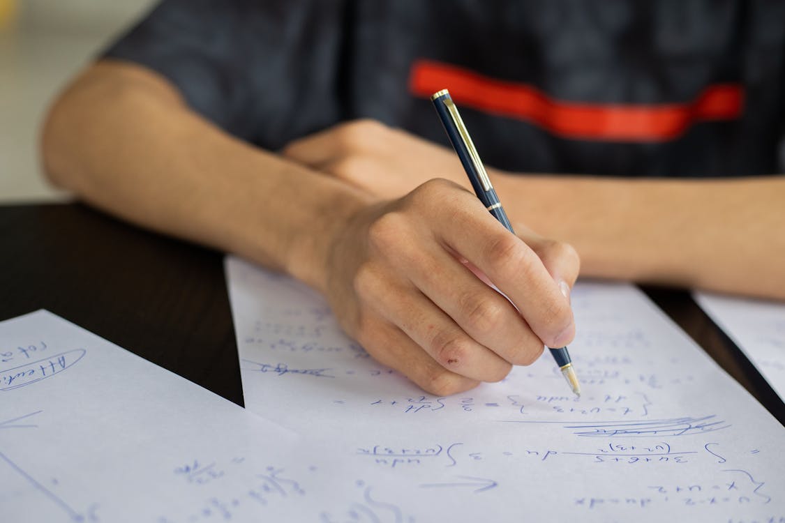 Free Unrecognizable smart male student with pen  taking notes while solving equations at table during lesson in classroom on blurred background Stock Photo