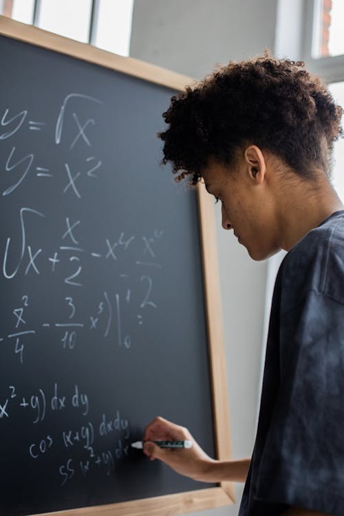 Side view of African American learner making math calculations on blackboard during lesson in classroom