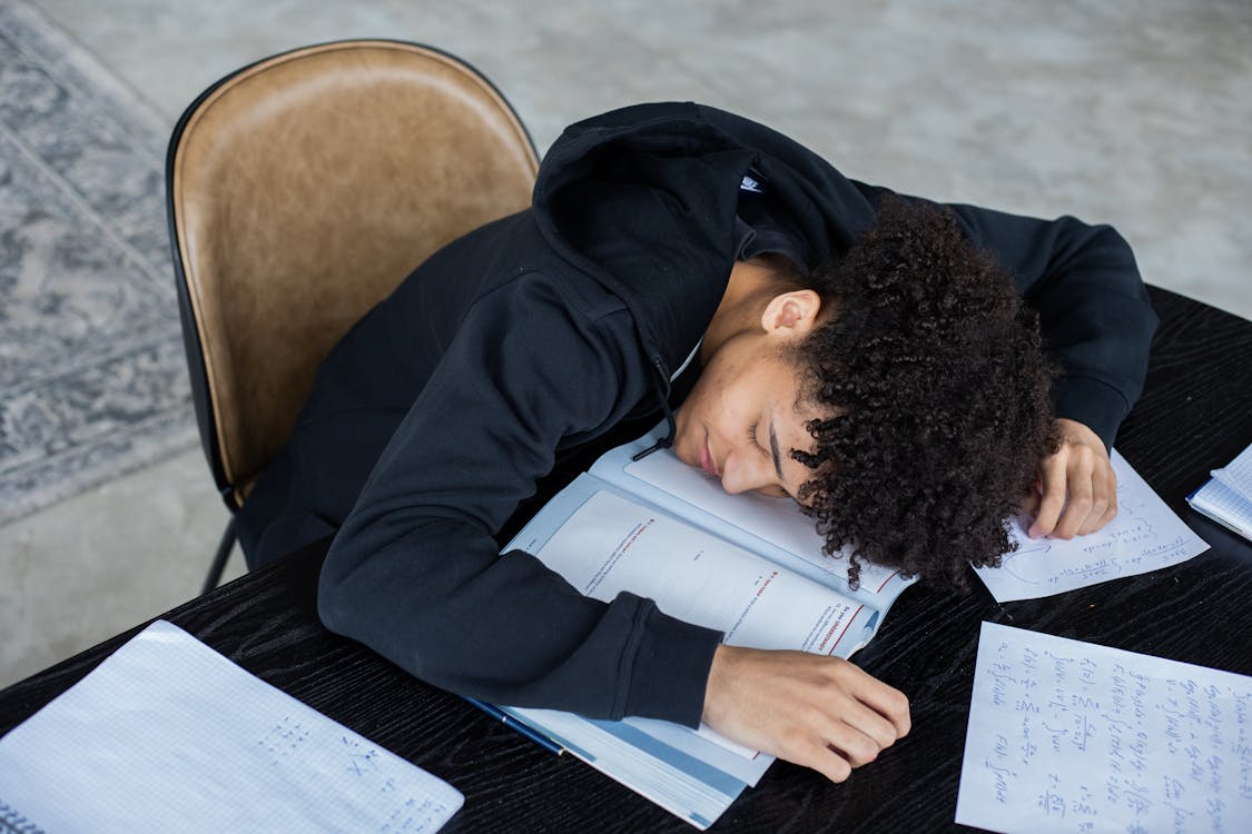 High angle of exhausted African American student resting on opened textbook and papers while preparing for exam