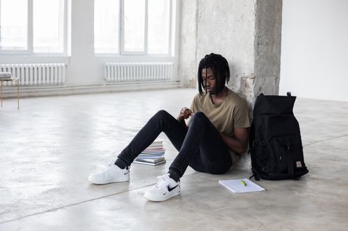Free Full body of upset African American man sitting among textbooks and notebook near backpack during break in exam preparation Stock Photo