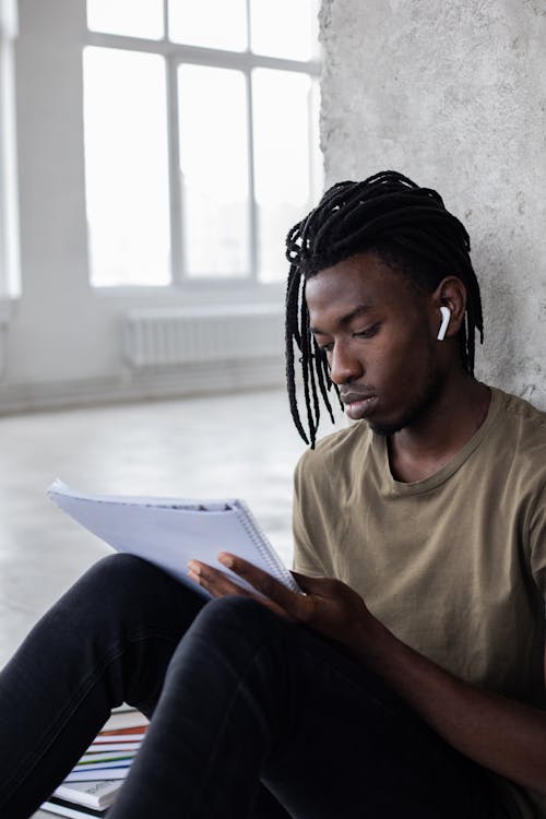 Focused African American male in wireless earbuds reading notes in notebook while getting ready for university assignment