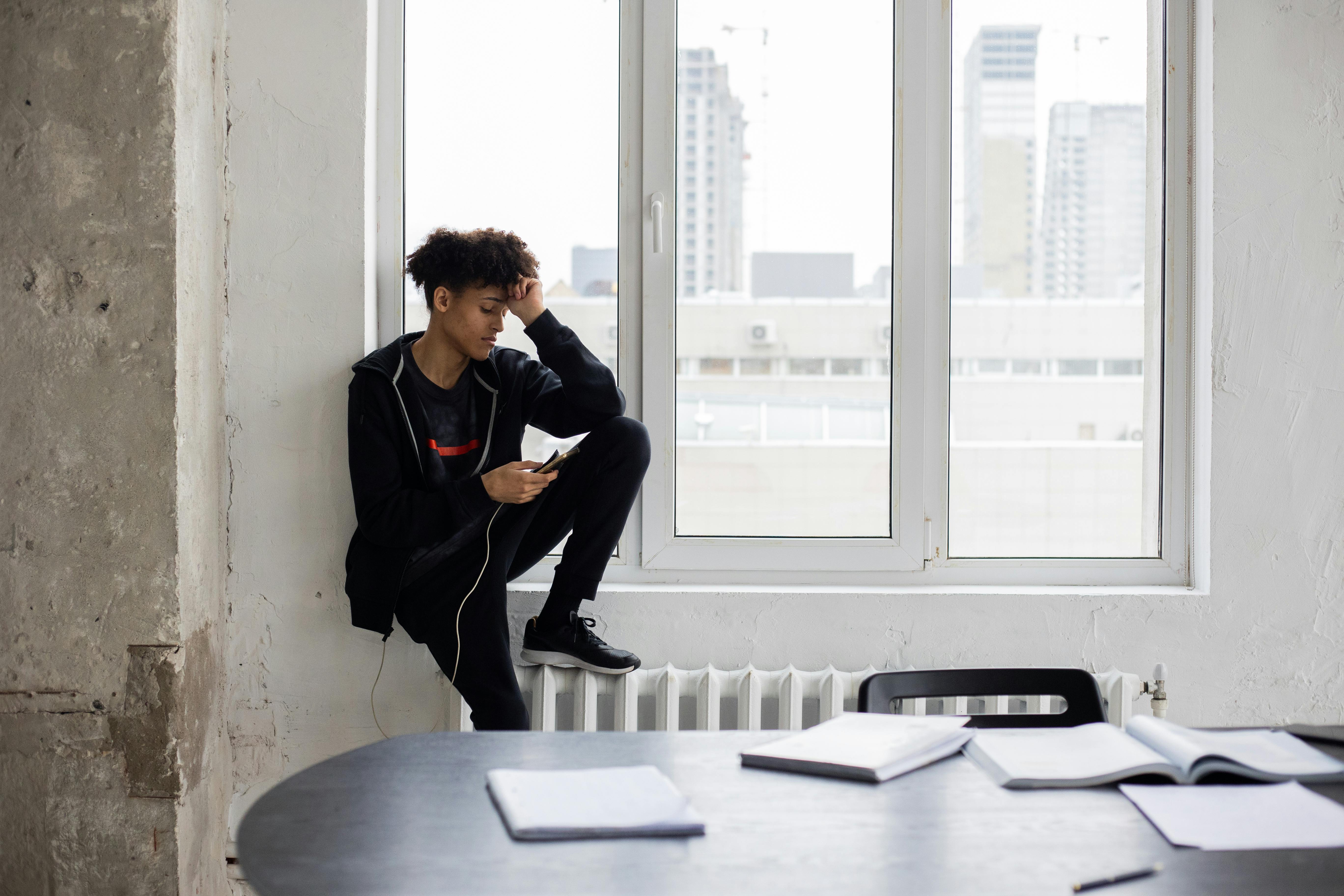 serious young black man browsing smartphone sitting on windowsill