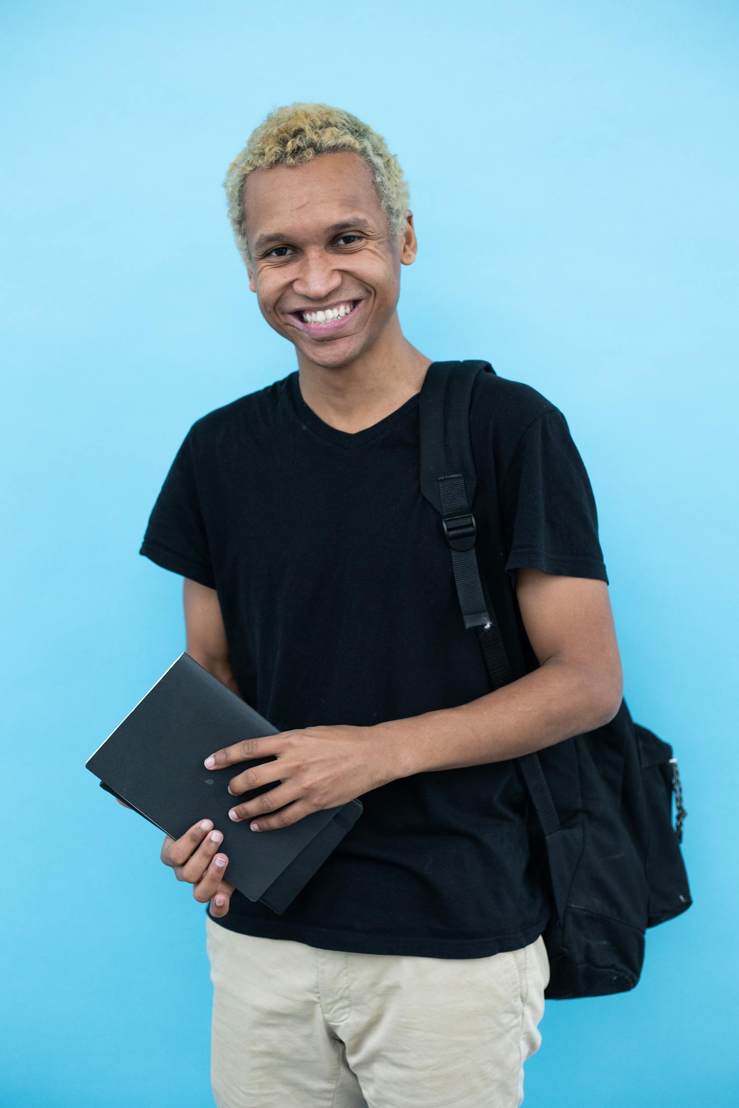 Smiling young man holding a book and wearing a backpack against a blue background.