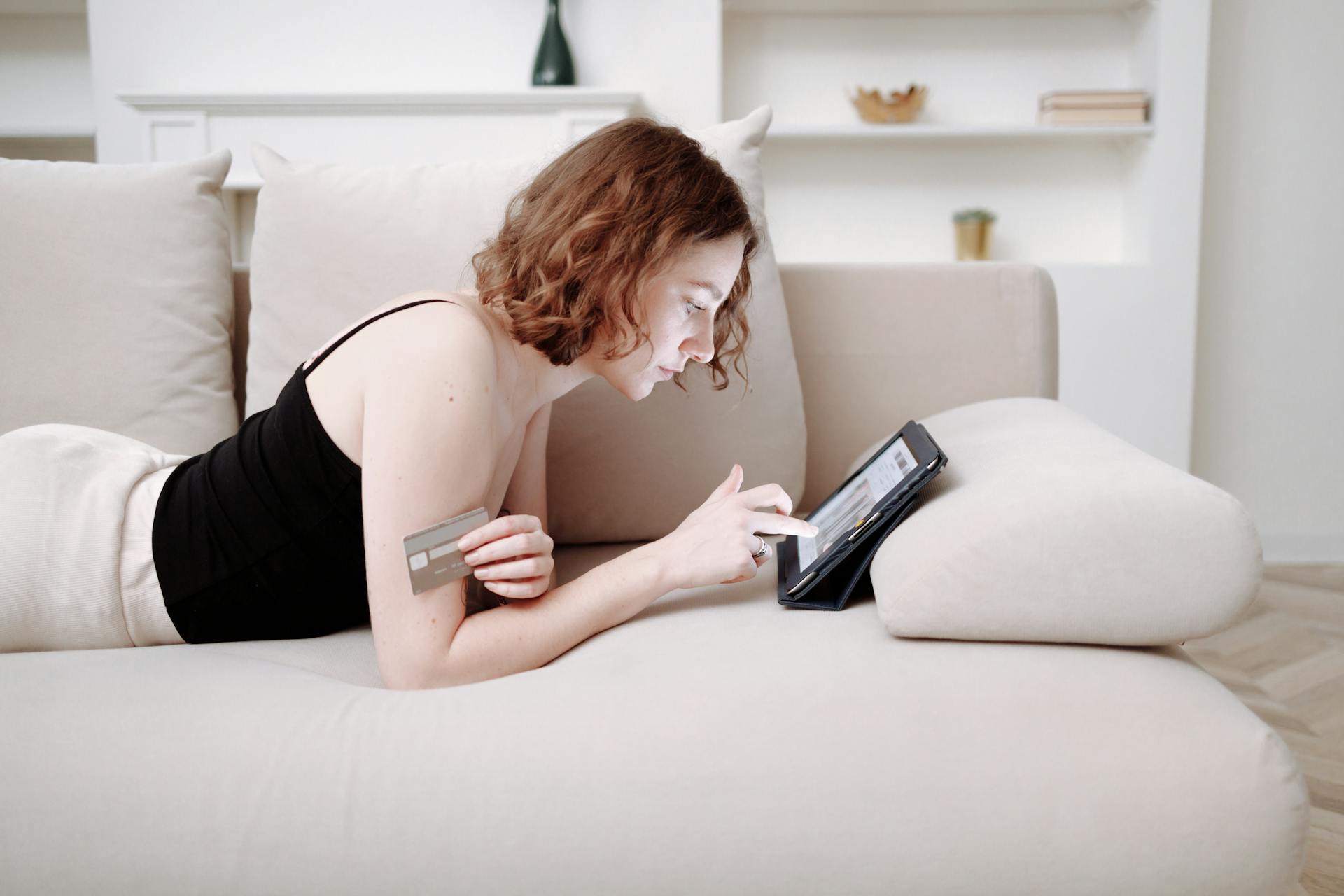 A woman shopping online comfortably on her couch using a credit card and tablet.