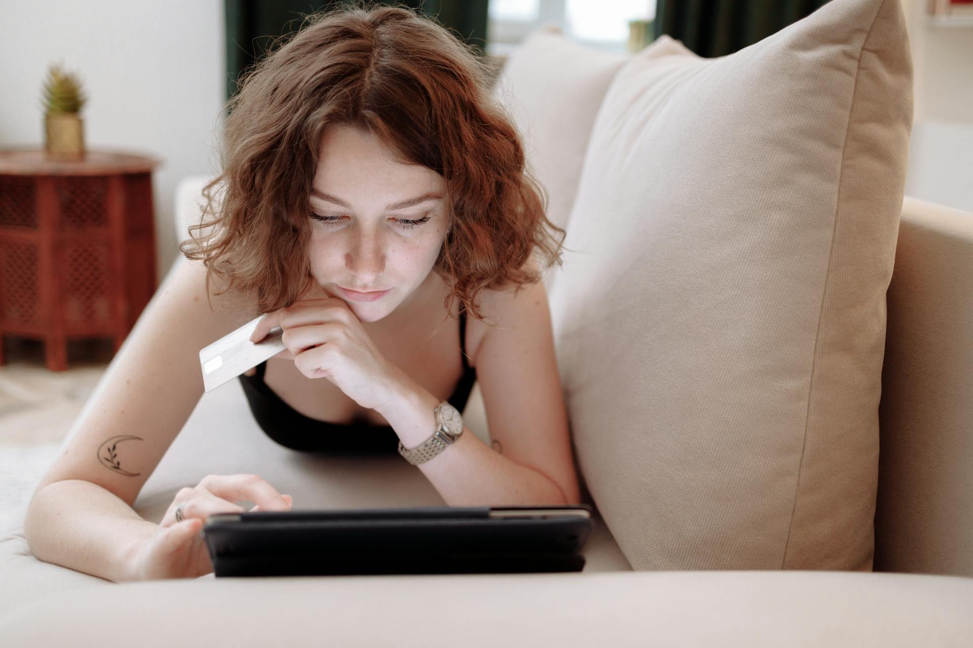 Young woman using a tablet and credit card for online shopping, lying on a couch indoors.