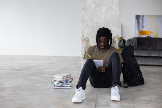 Full body of African American male student doing homework assignment in spacious room with pile of textbooks and backpack