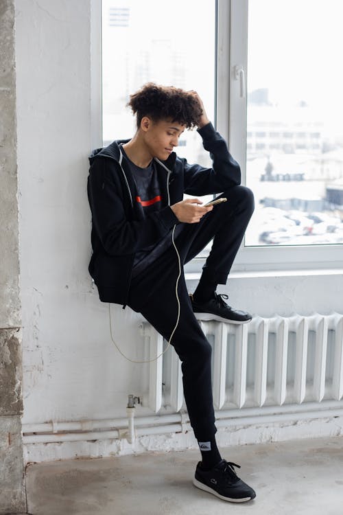 Free Full body of African American male with curly hair sitting near window and browsing charging mobile phone Stock Photo