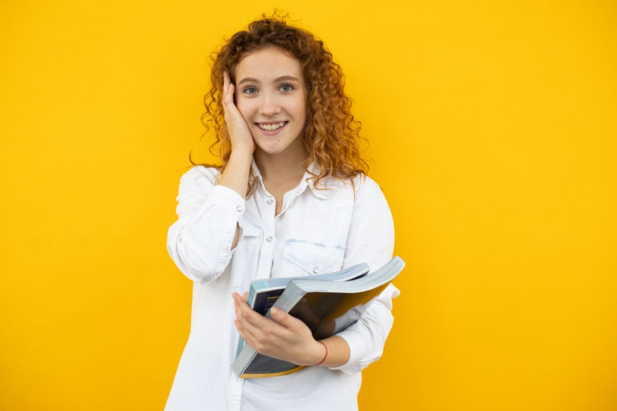 Surprised female student with curly hair standing with textbooks and smiling at camera against yellow background