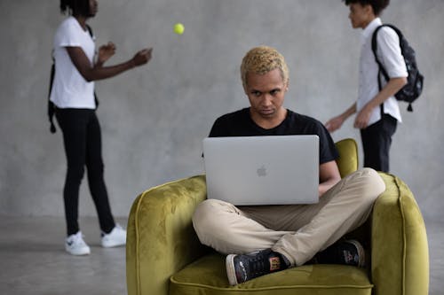 Concentrated African American learner sitting in chair and browsing netbook for studies against classmates playing with ball