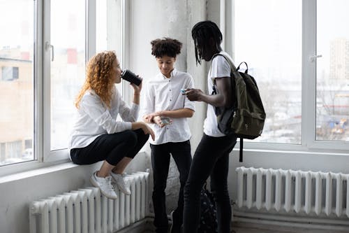 Free Group of diverse students drinking takeaway coffee and browsing smartphones while standing near window in classroom before lesson in college Stock Photo