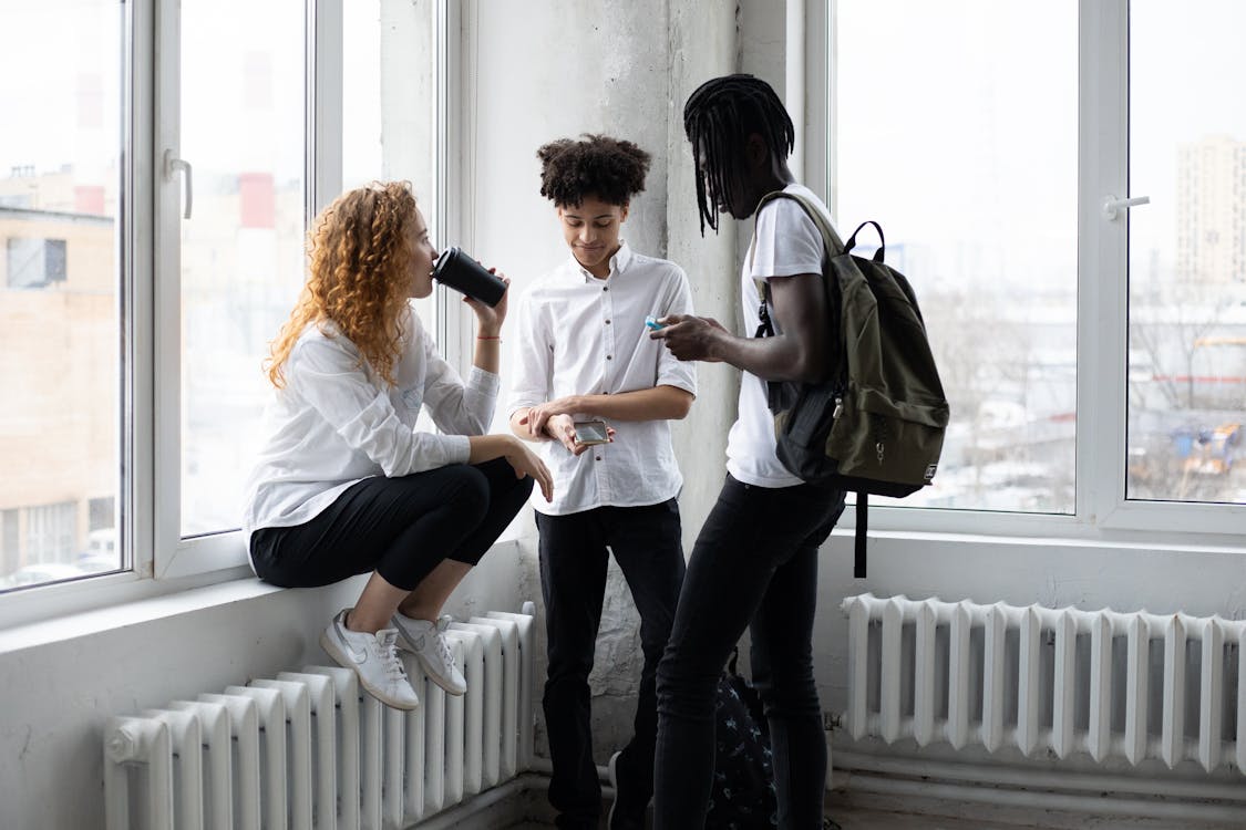 Group of diverse students drinking takeaway coffee and browsing smartphones while standing near window in classroom before lesson in college