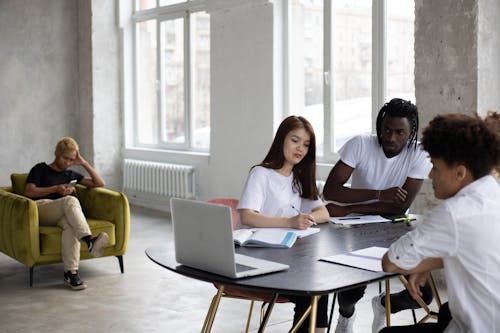Group of young multiethnic coworkers in casual clothes gathering at table with laptops and papers and discussing project details in light spacious workspace