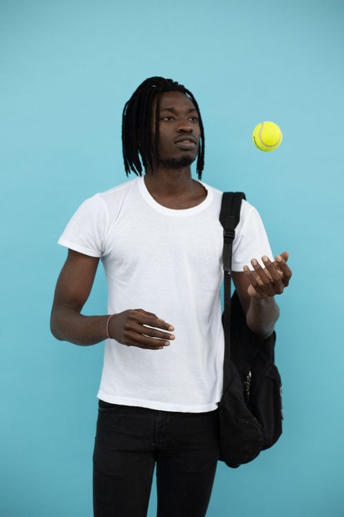 Free Focused young African American male in casual wear with backpack throwing tennis ball against blue wall in studio Stock Photo