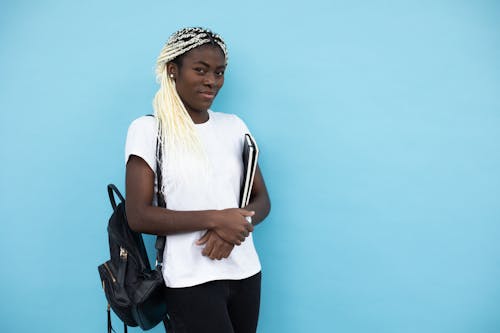 Content young African American female student with dyed white braids standing with books and backpack against blue background and looking at camera