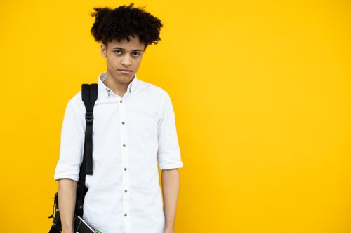 Free Calm young African American male student with Afro hairstyle wearing white shirt with backpack standing on yellow background and looking at camera Stock Photo