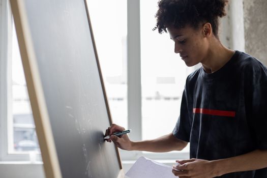 Crop focused young African American male in casual outfit writing on blackboard in light classroom or office