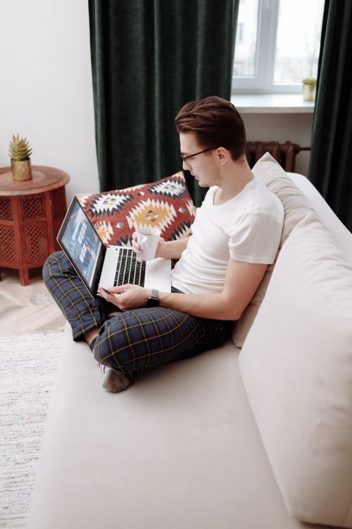 Free Young man sipping coffee and browsing laptop on couch at home. Stock Photo