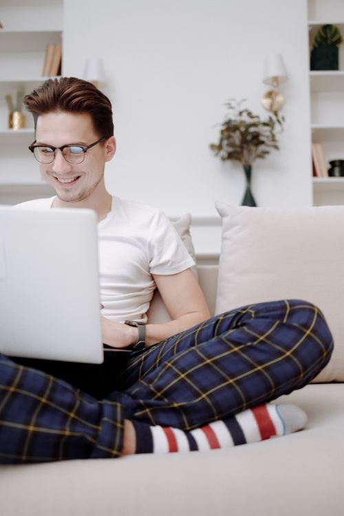 Free Smiling young man in glasses sitting cross-legged on a couch, using a laptop indoors. Stock Photo